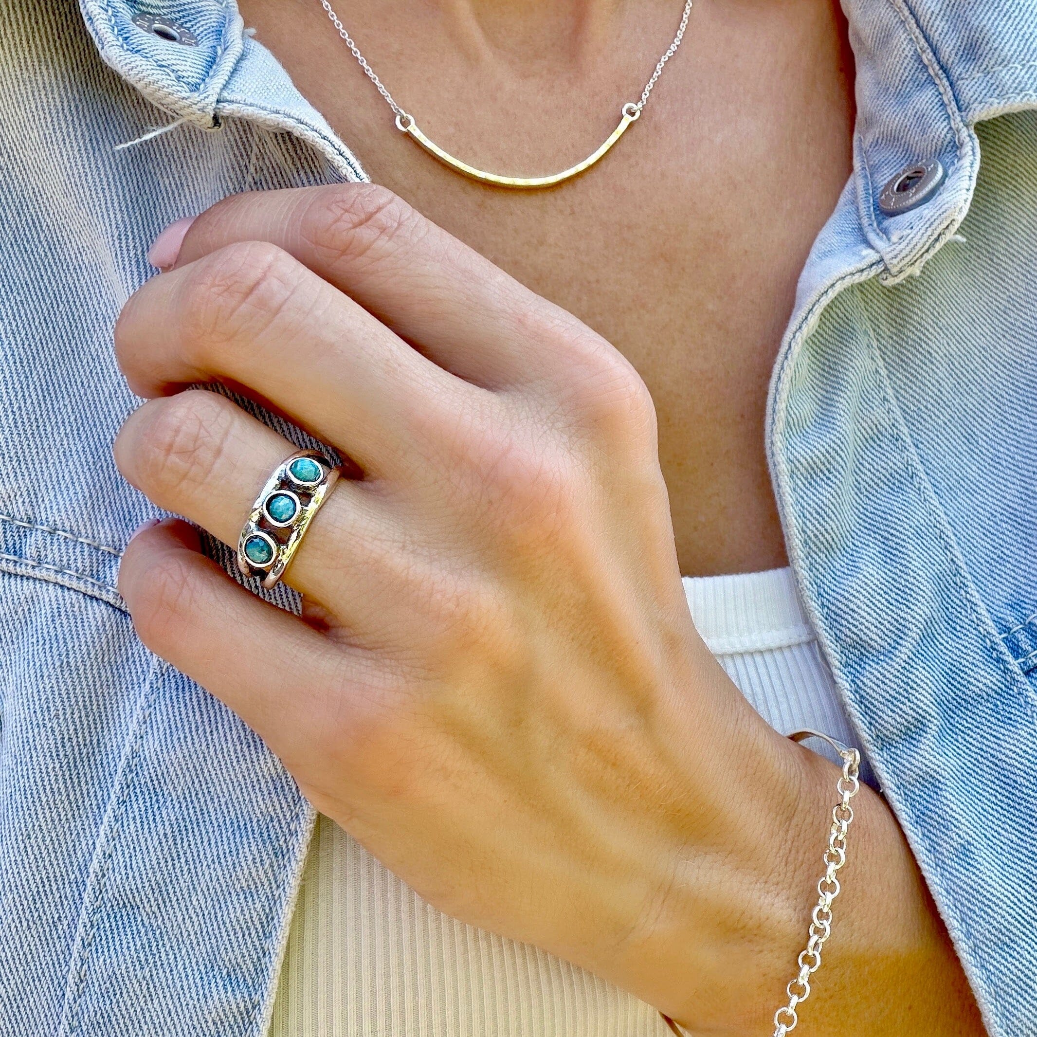 A close-up shot of a silver band ring with three round turquoise-blue stones, styled with a silver necklace and bracelet.