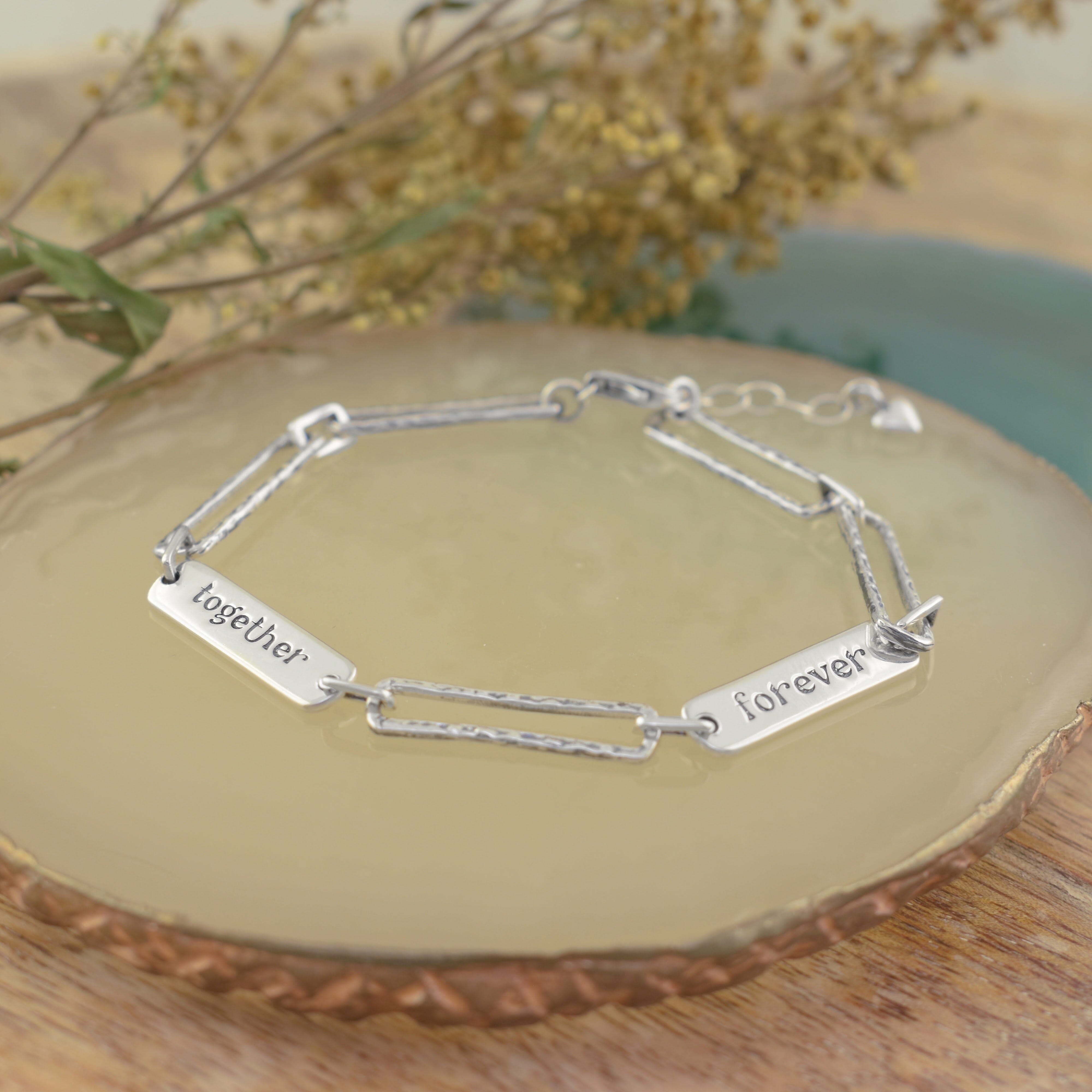 A sterling silver chain bracelet with rectangular links, one engraved with 'together' and another with 'forever', and a small heart charm hanging from the end of the extender
