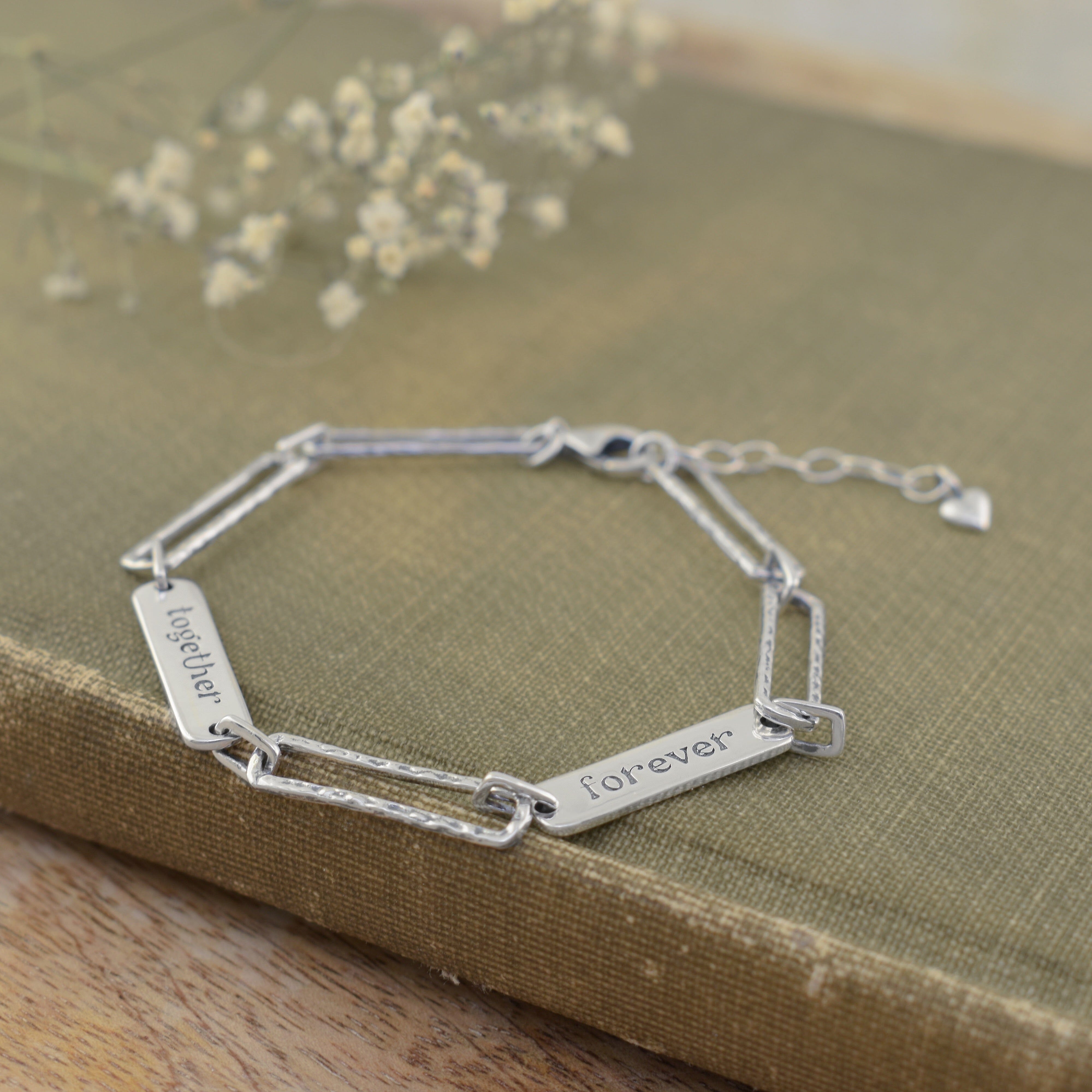 A sterling silver link bracelet with two engraved rectangular plates reading 'together' and 'forever', with a small heart charm hanging from the extender