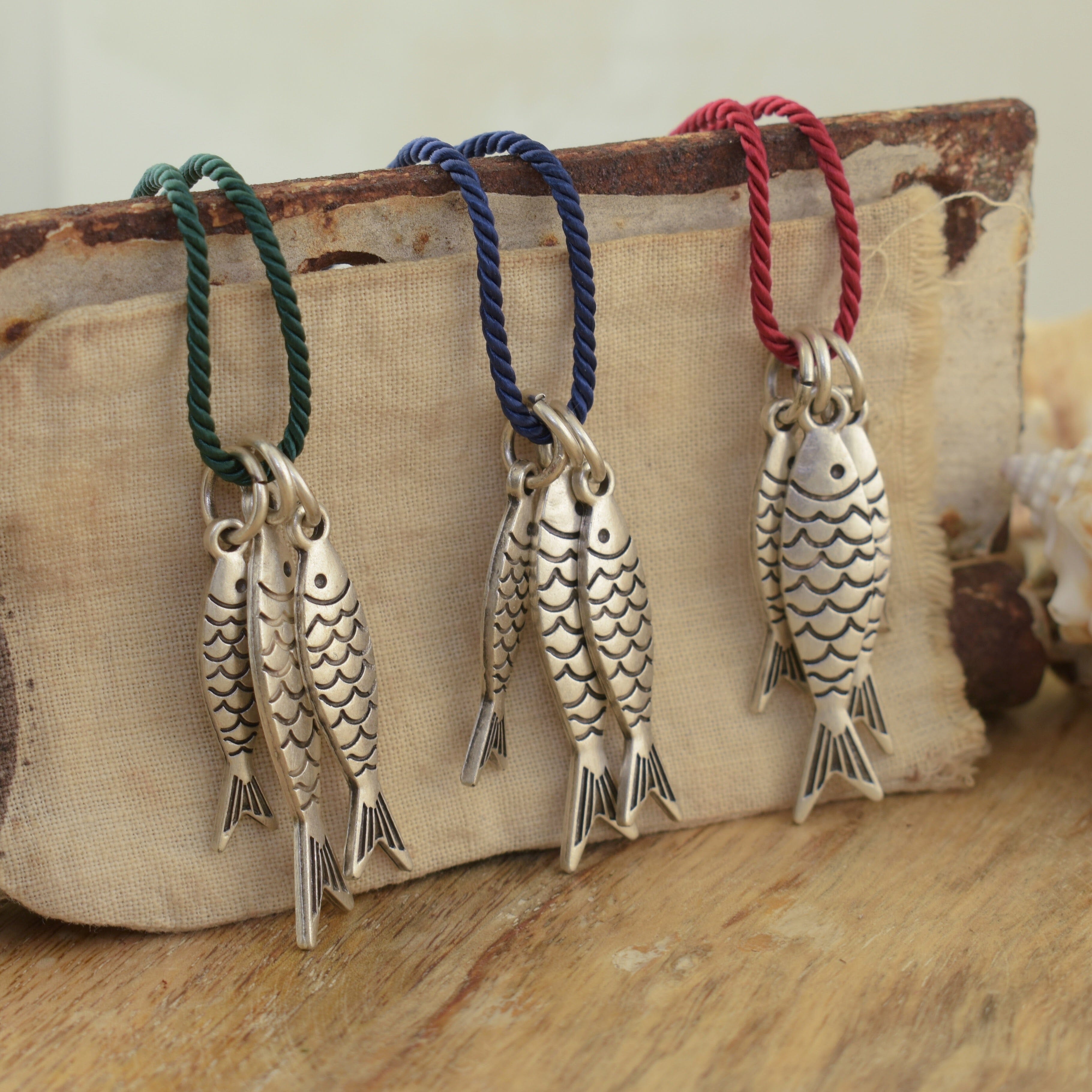 Three fish-shaped pendant necklaces with detailed scales hang from braided cords in green, blue, and red. 