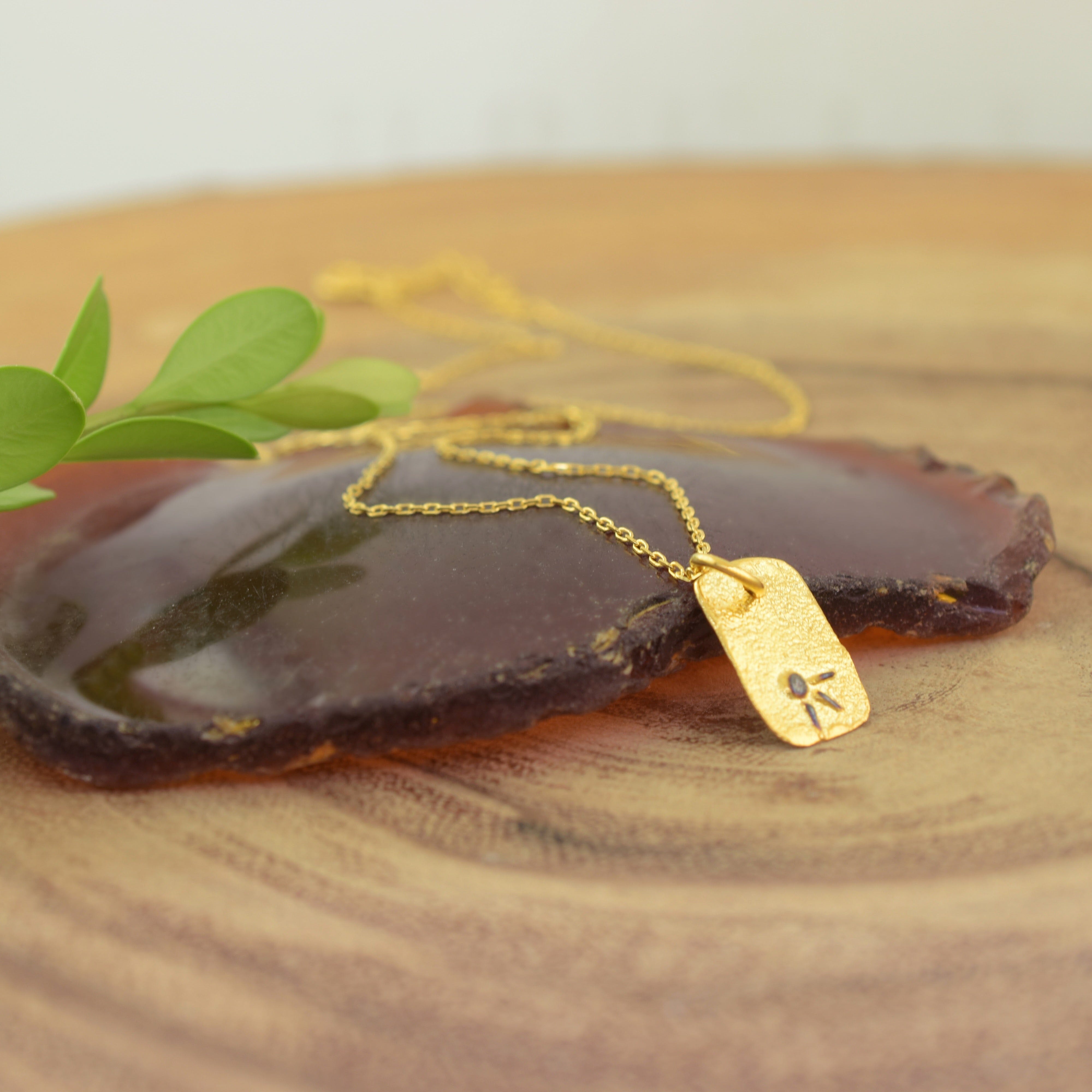 A gold necklace with a rectangular pendant, featuring a small sun burst symbol