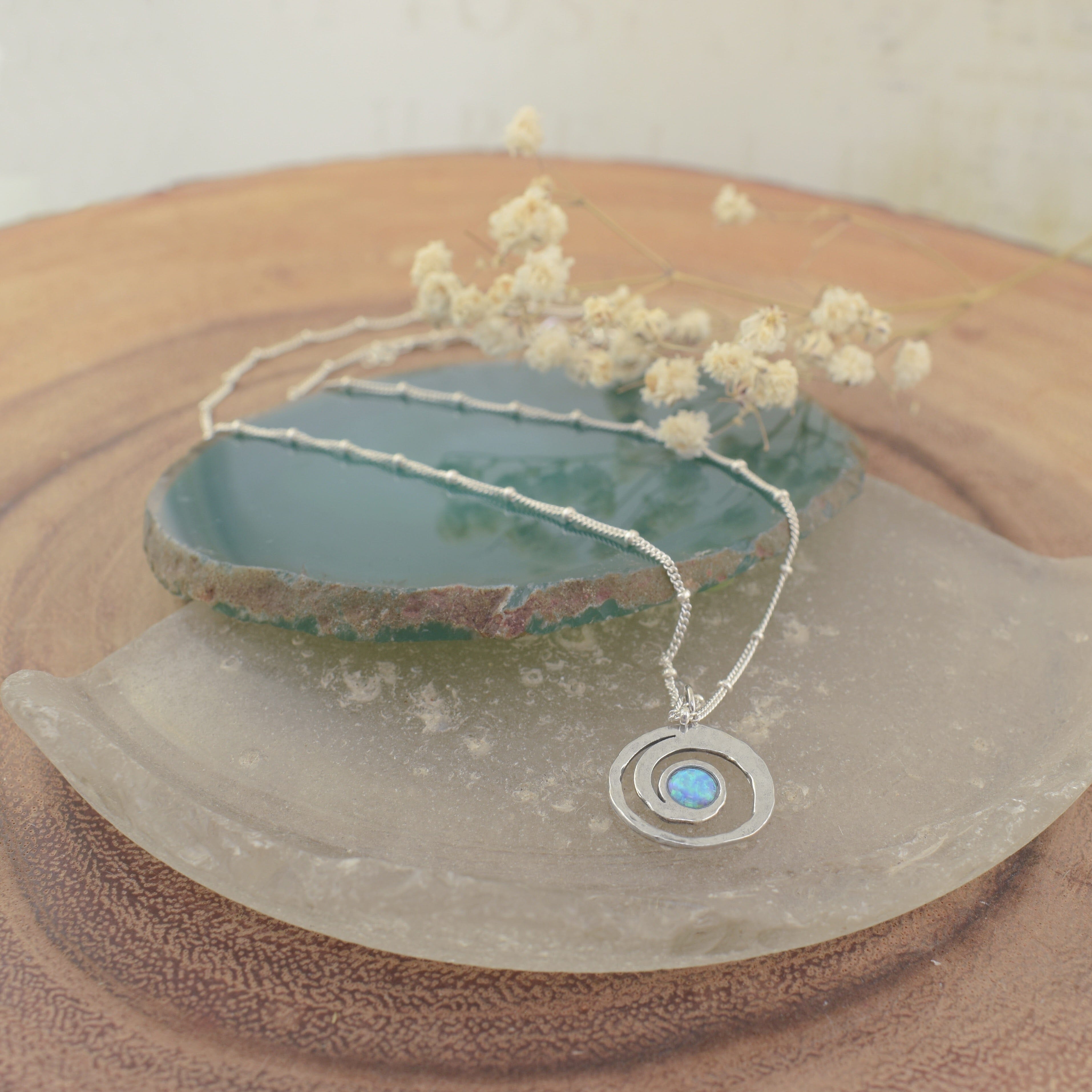 A sterling silver necklace with small silver beads spaced along the chain, featuring a round spiral pendant with an opal stone resting in the middle