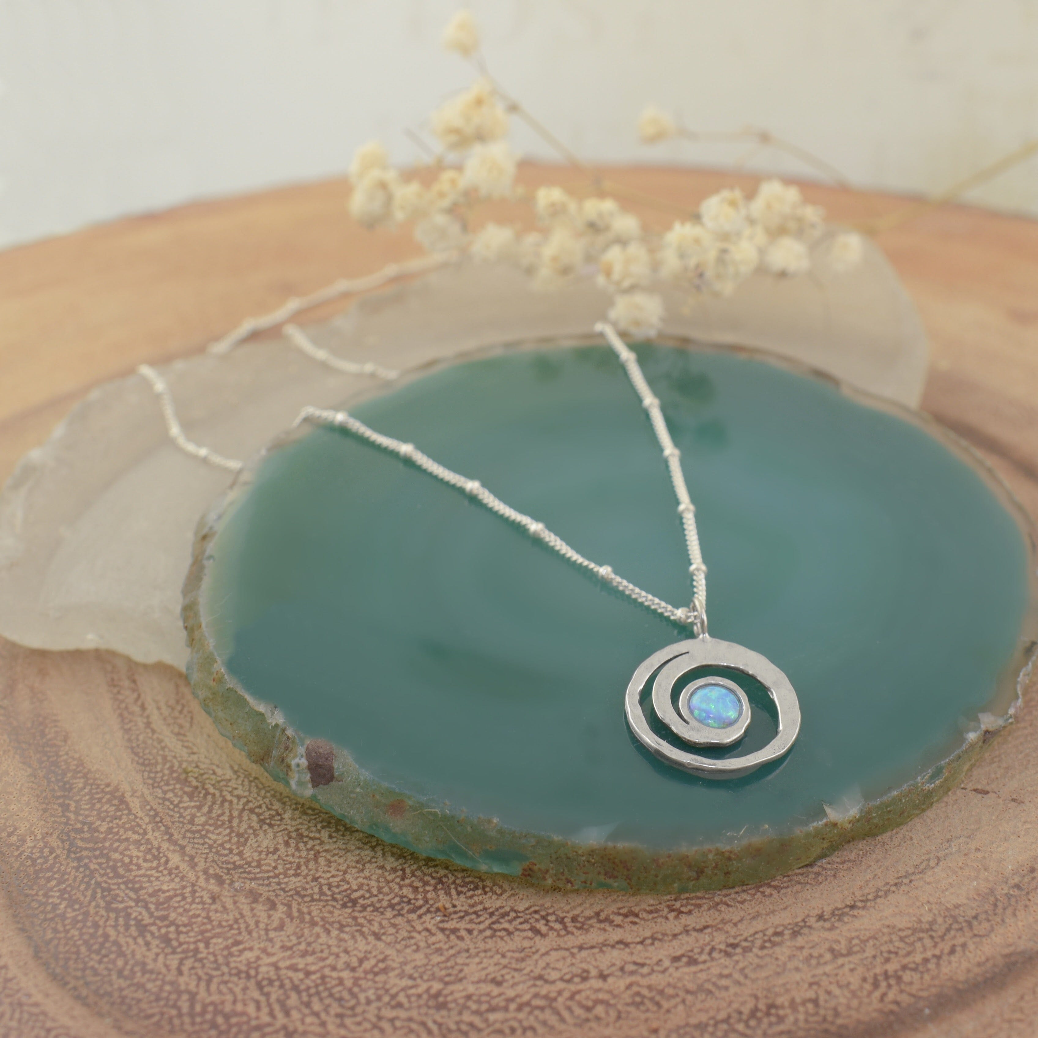 A sterling silver necklace with small silver beads spaced along the chain, featuring a round spiral pendant with an opal stone resting in the middle