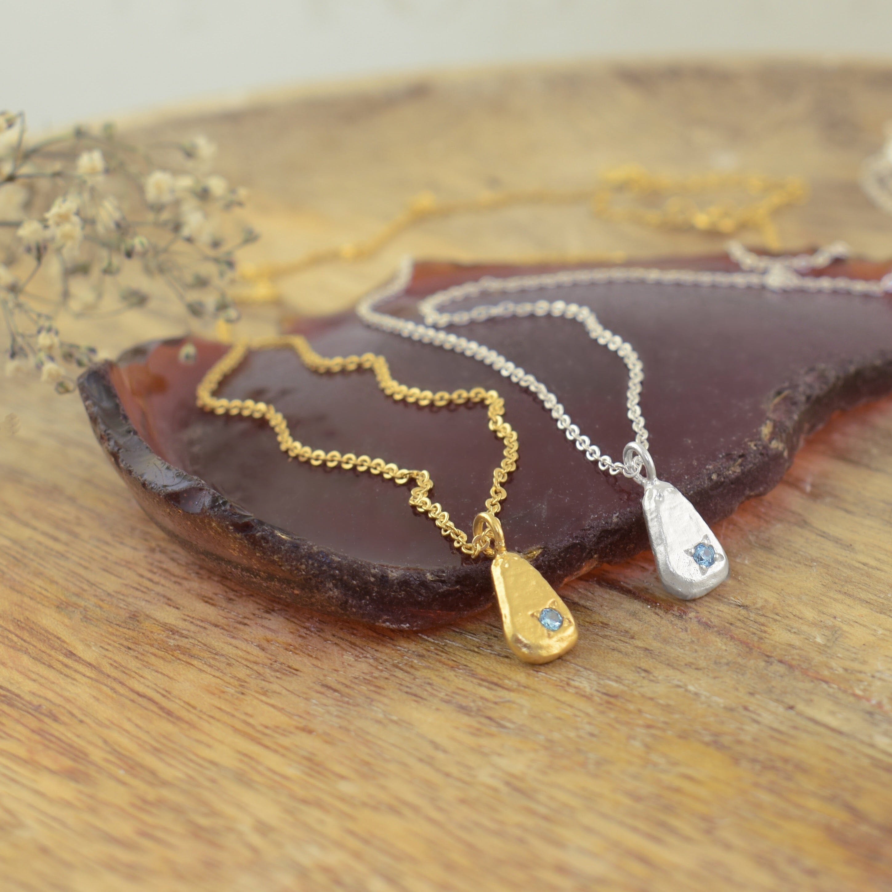 Two teardrop-shaped pendant necklaces with small light blue gemstones, one in gold and one in silver
