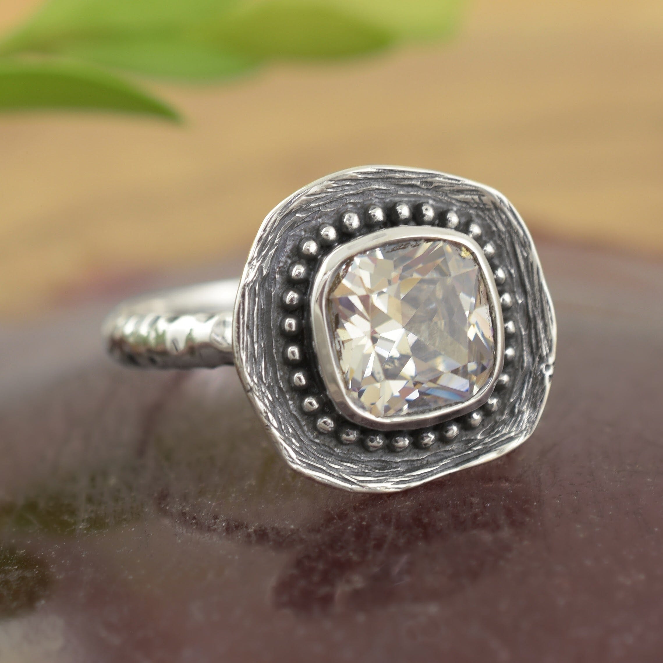 A sterling silver ring, featuring a square cut clear CZ, with small silver beads framing the stone and a textured square base for it to sit on