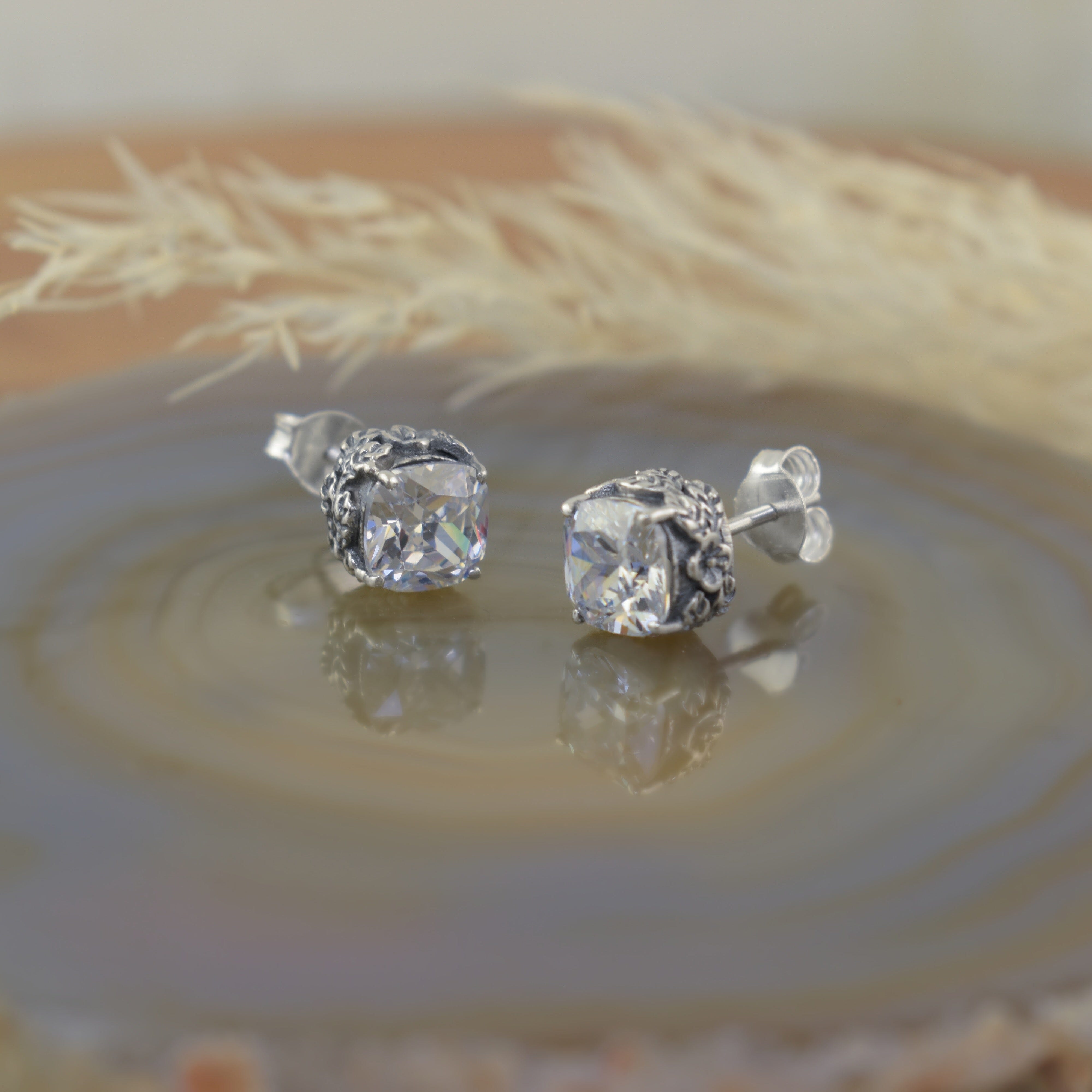 A small pair of sterling silver stud earrings with a square-cut CZ stone and vine-like details along the sides