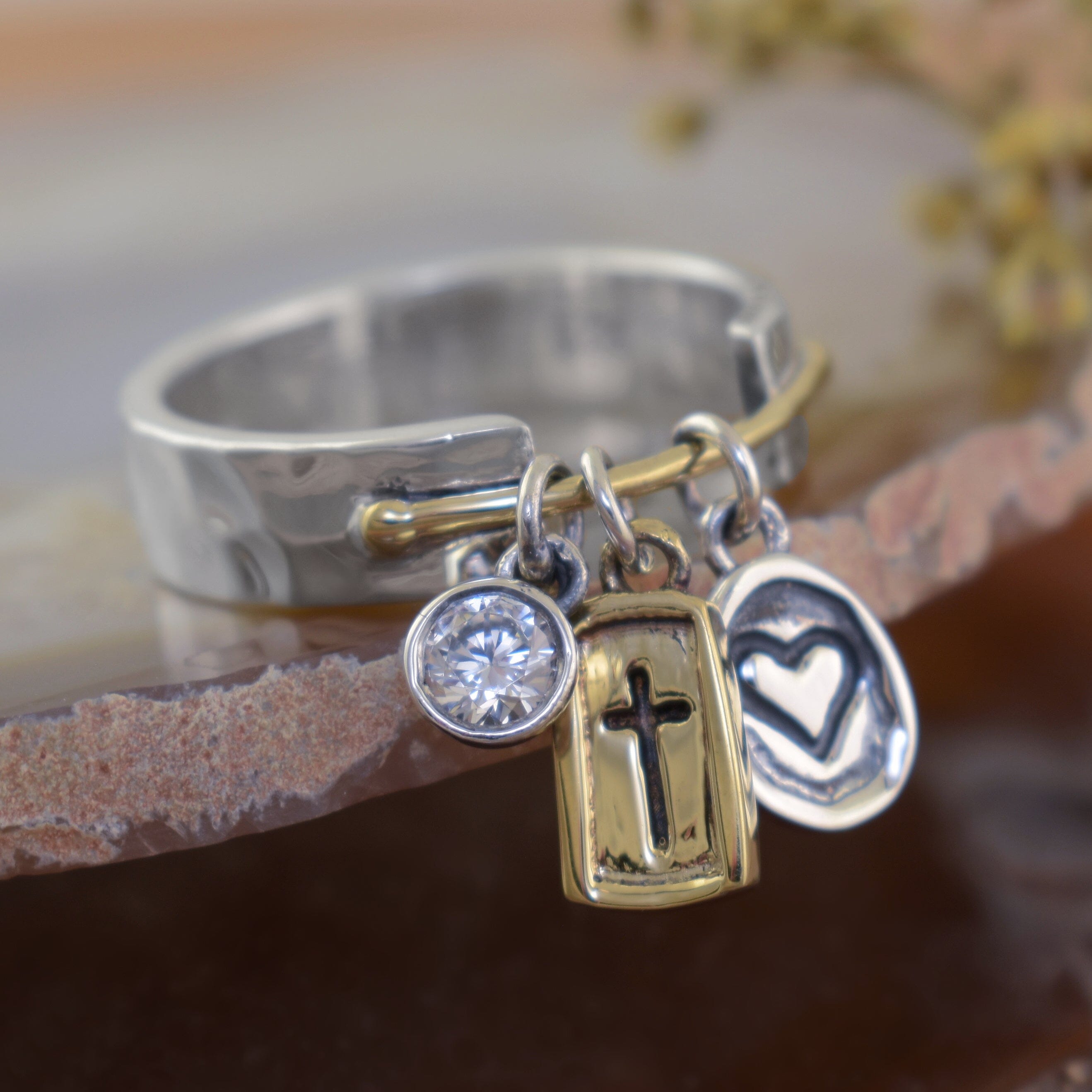 a sterling silver ring with a gold bar connecting 3 individual pendants, one pendant is a clear CZ, one is a gold rectangle engraved with a cross, one is a silver circle with an engraved heart