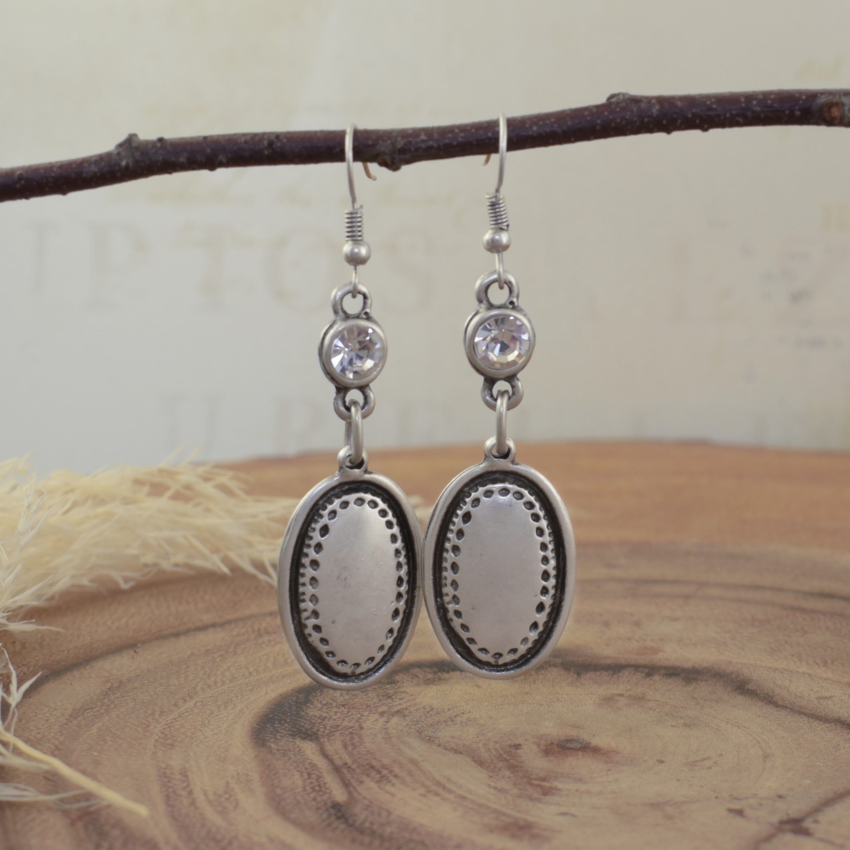 A pair of dangly silver-plated zamak earrings, featuring a round clear CZ stone and large oval with textured detailing hanging from the hooks