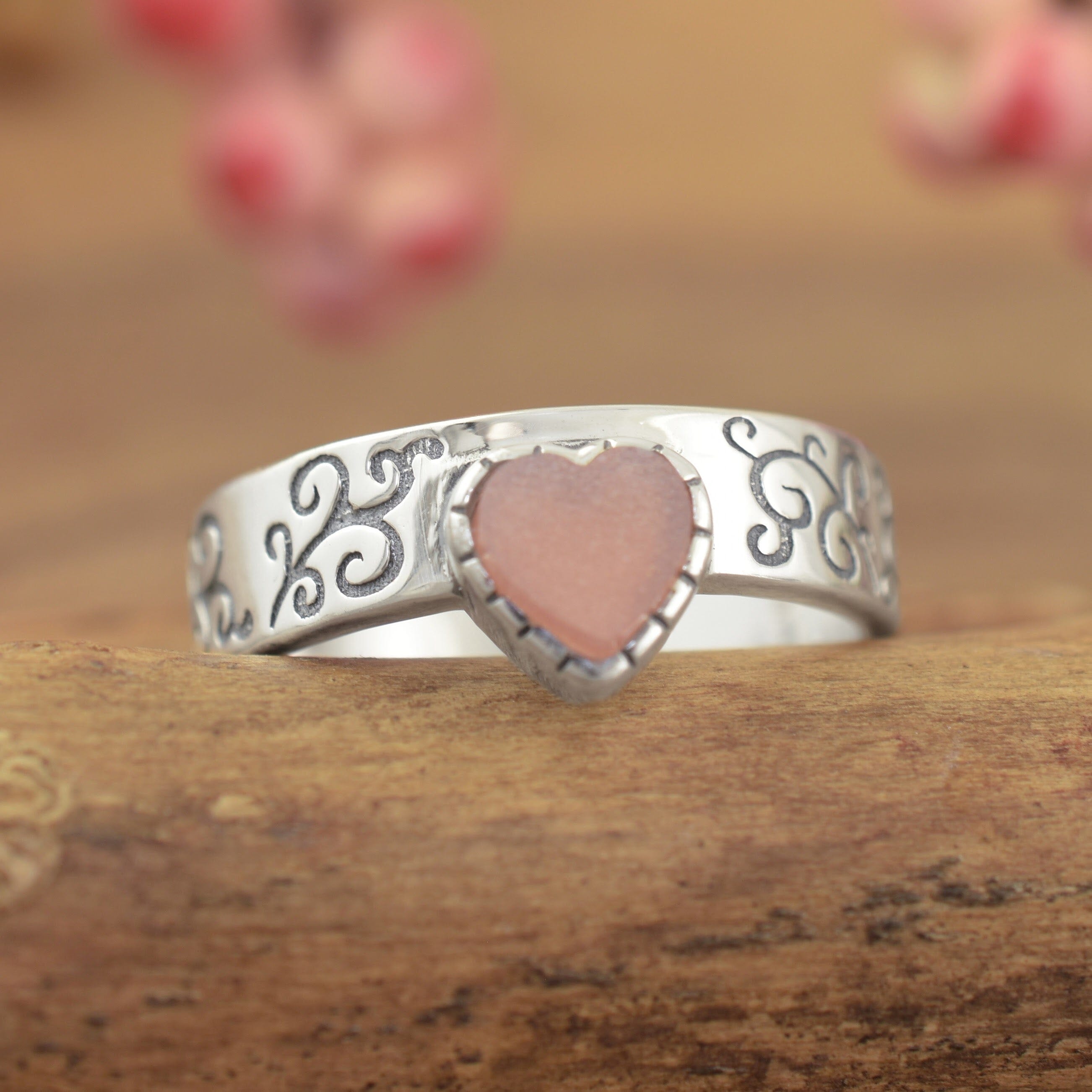 A sterling silver ring featuring a pale salmon-colored heart, with engraved filigree along the high-polished band