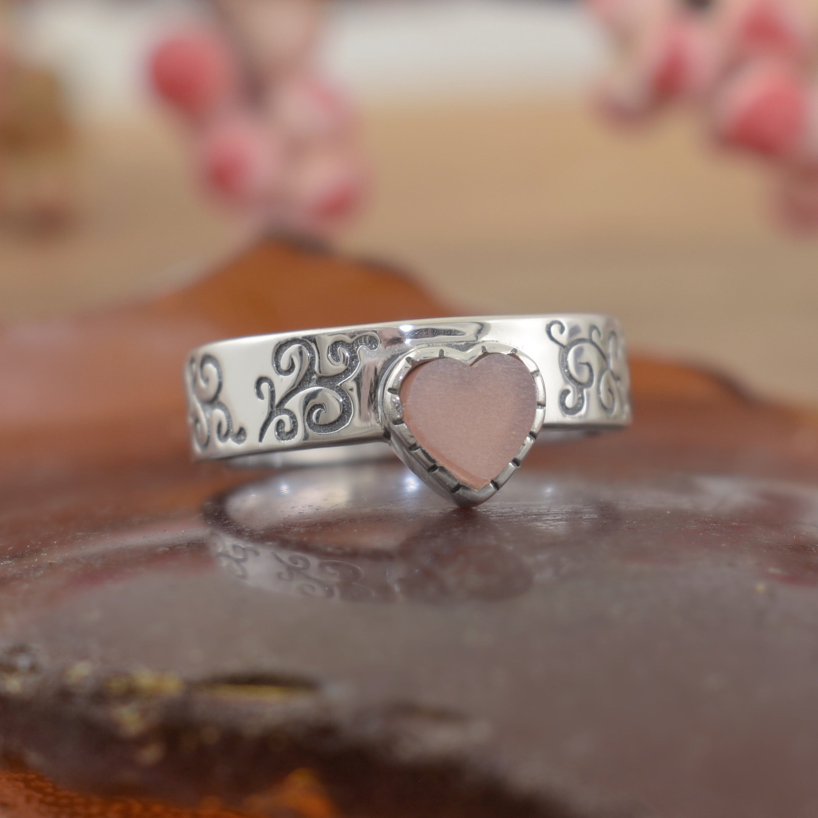 A sterling silver ring with engraved filigree along the high polished band, featuring a pale pink heart in the center