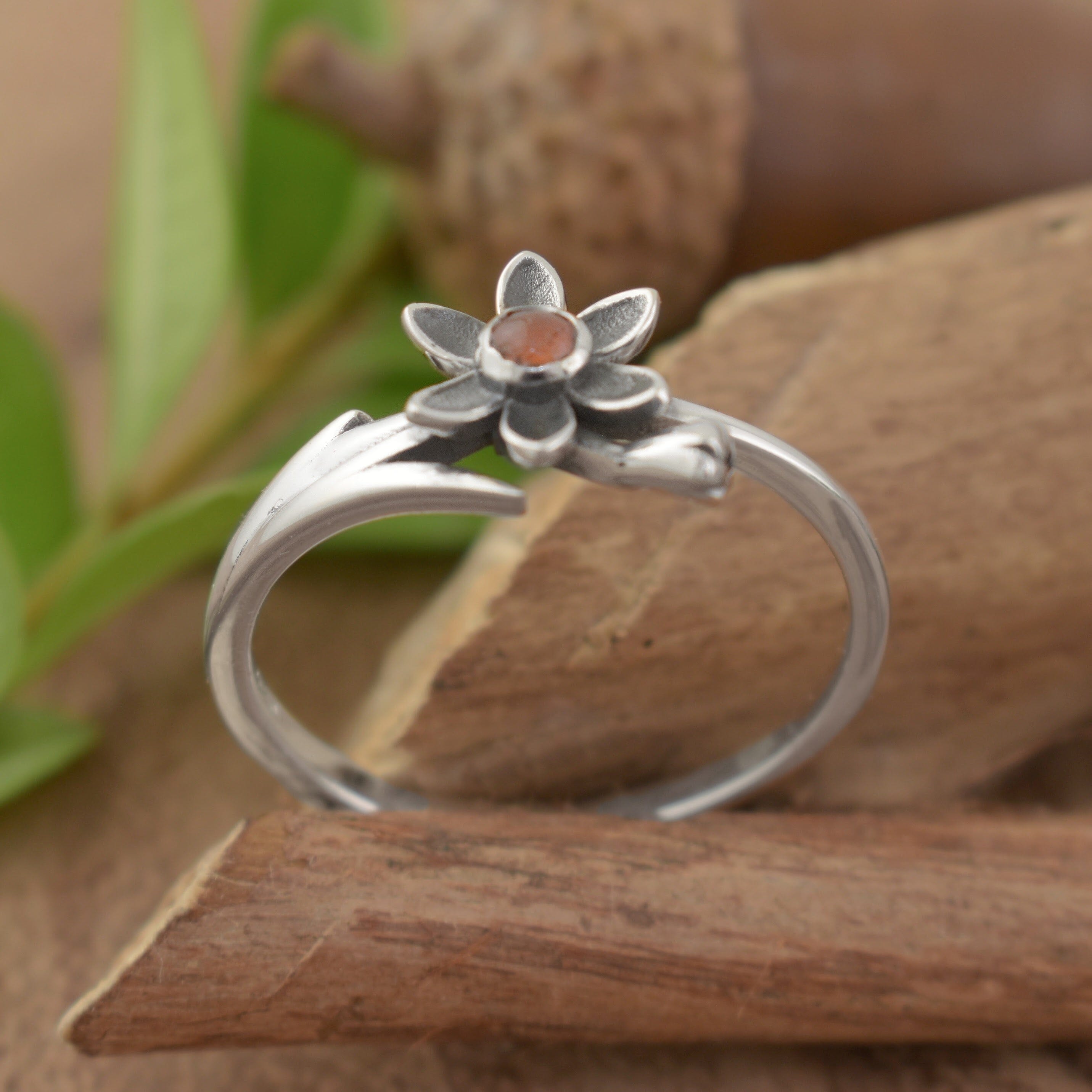 A small sterling silver ring with an orange stone in the center of a silver flower and an organic flower stem design along the band