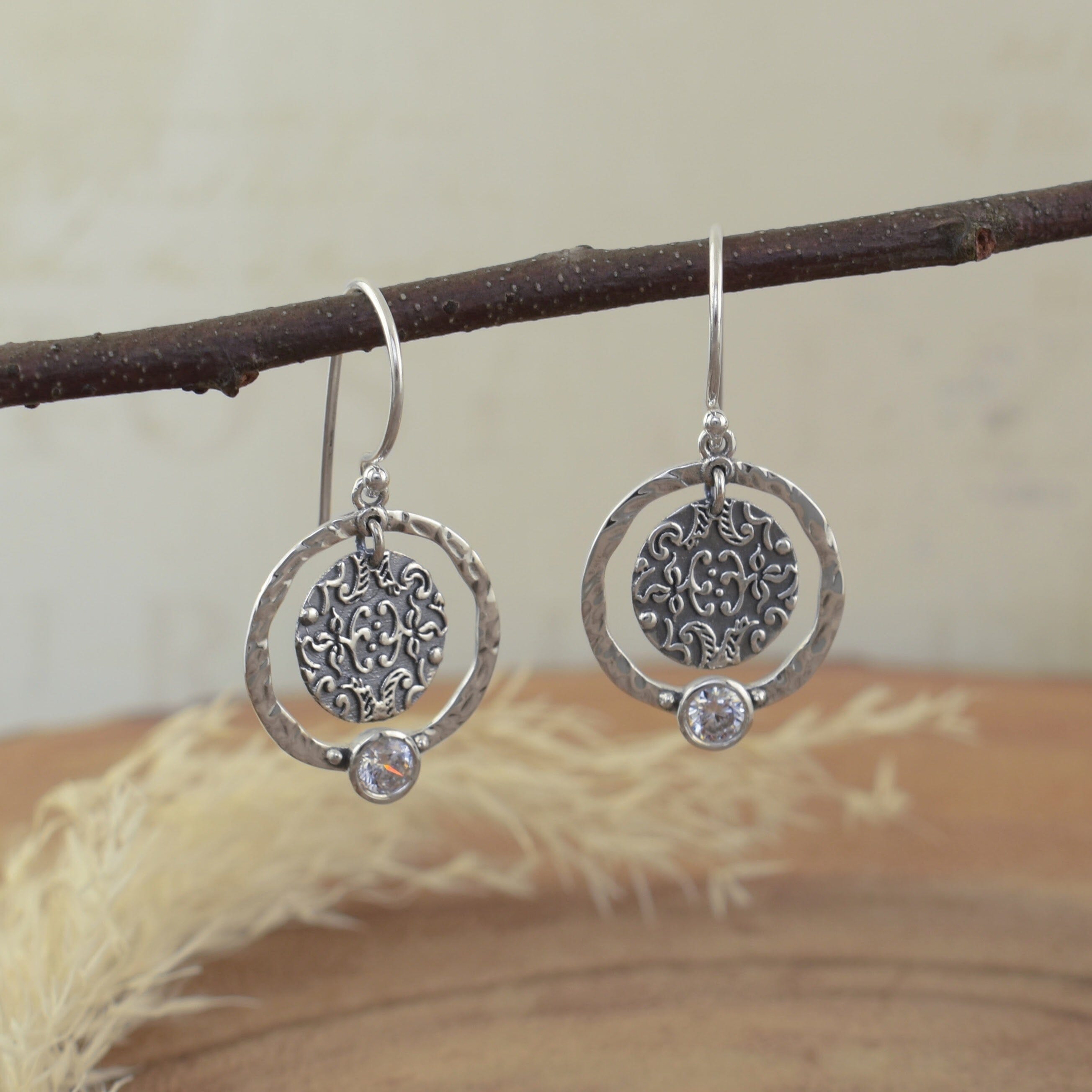 A pair of sterling silver earrings with a hammered ring and small engraved disc hanging in the middle with a small CZ stone at the bottom of the ring
