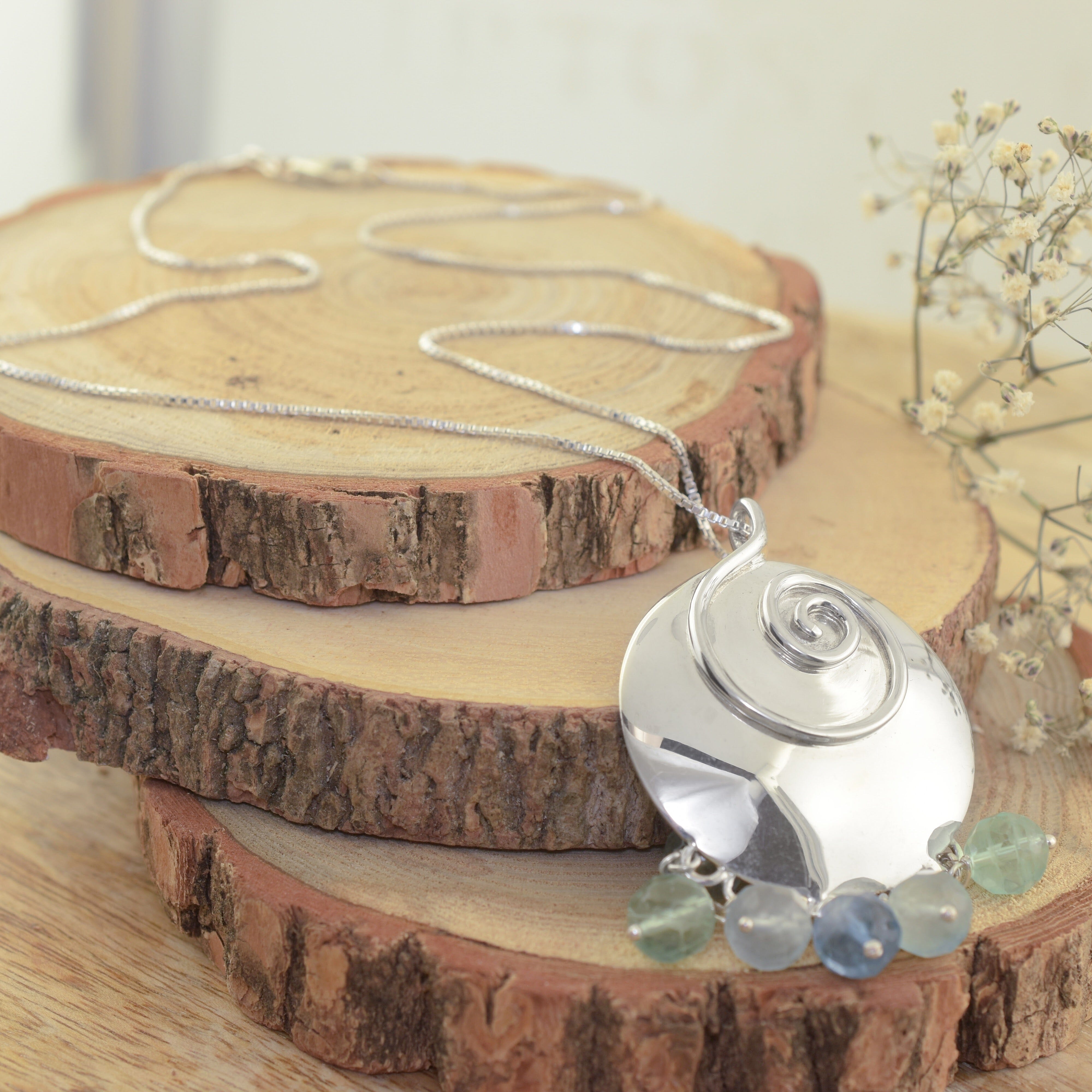 A sterling silver necklace featuring a large circle pendant, with a raised silver swirl design and pale green, blue and teal beads dangling from it