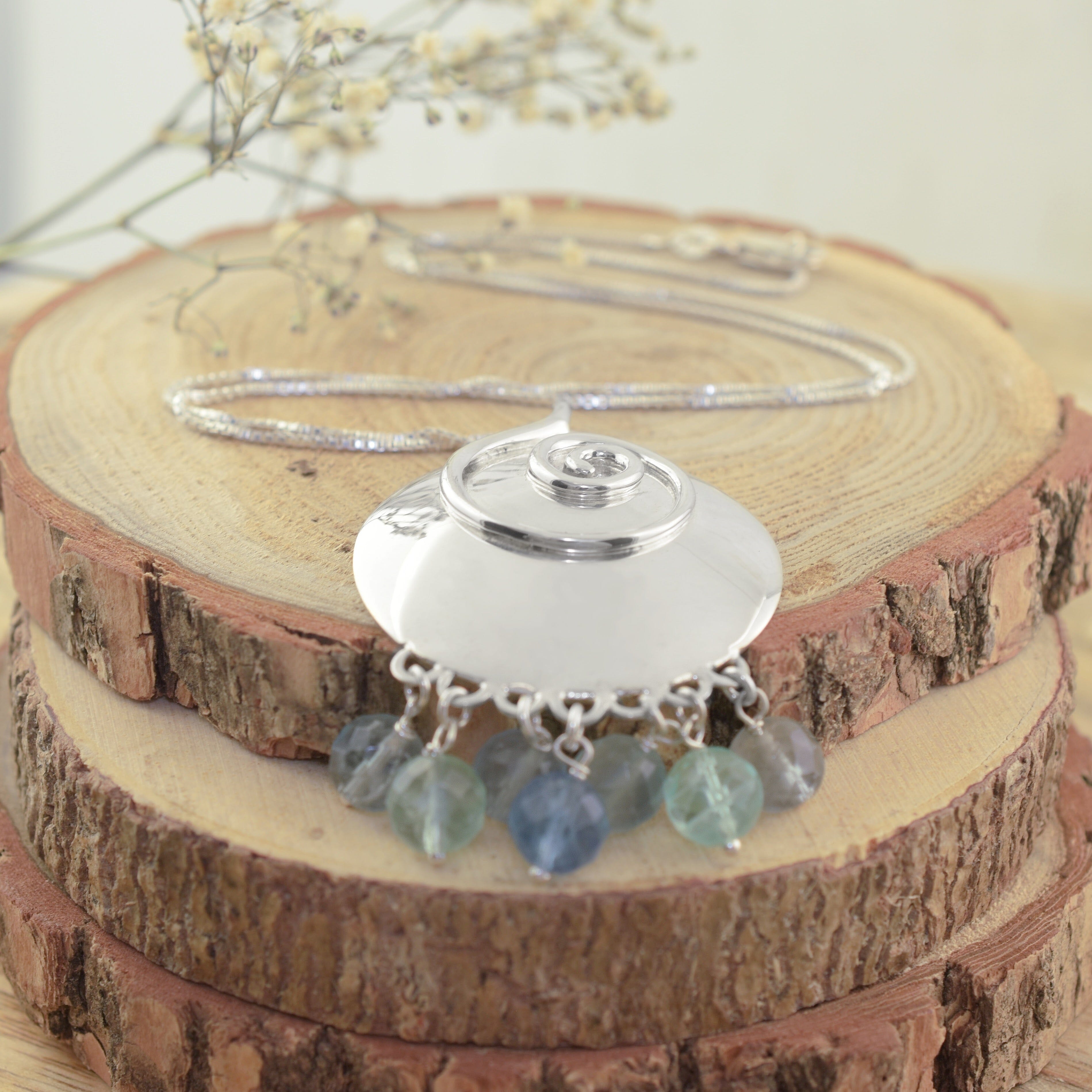 A sterling silver necklace featuring a large circle pendant, with a raised silver swirl design and pale green, blue and teal beads dangling from it