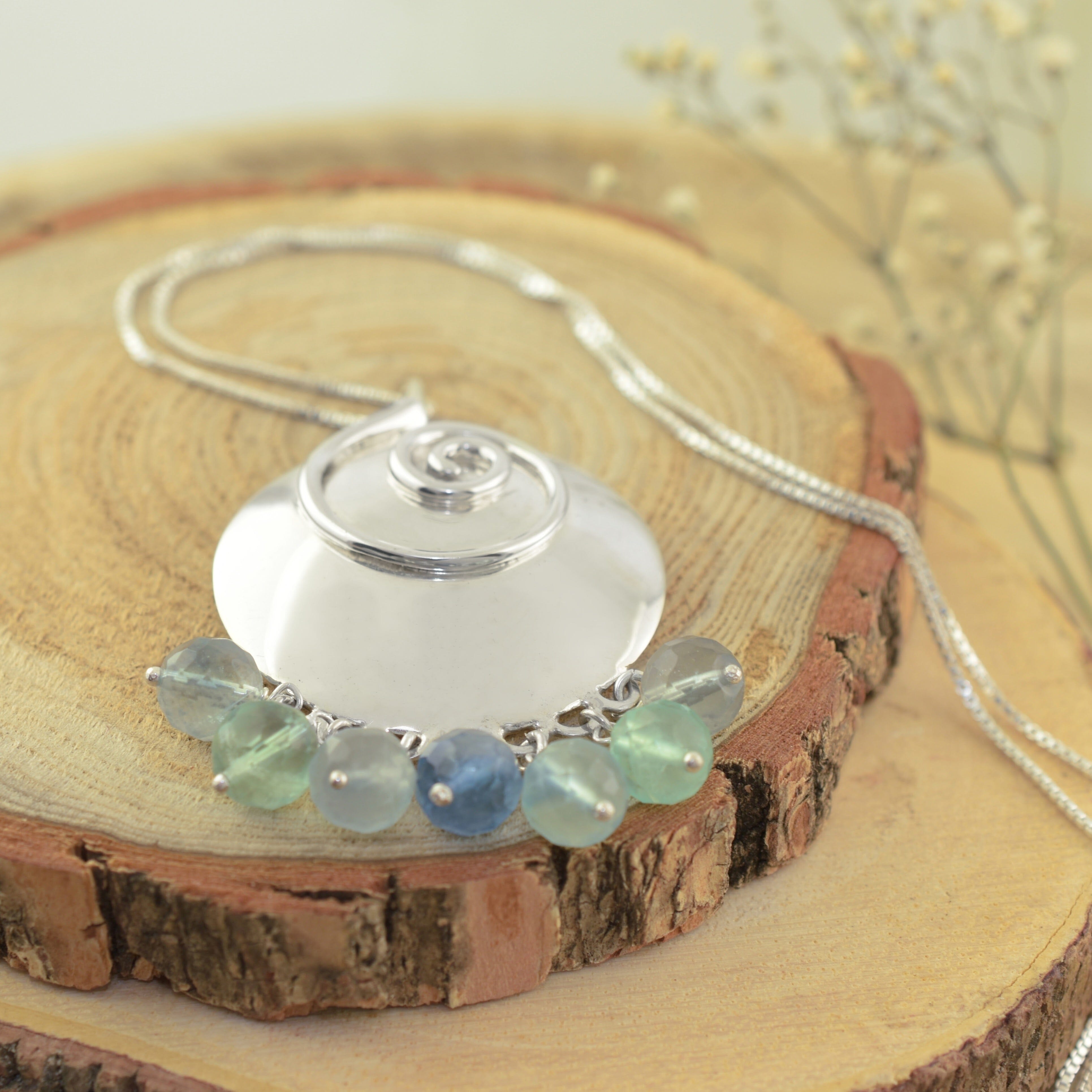 A sterling silver necklace featuring a large circle pendant, with a raised silver swirl design and pale green, blue and teal beads dangling from it